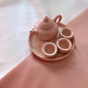 May include: A miniature tea set featuring a small teapot and three matching cups arranged on a round tray. The set is crafted from a smooth, glossy material with a marbled pattern in shades of soft pastel pink and white throughout the pieces.