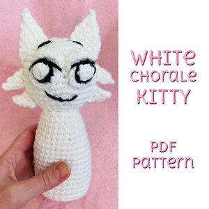 May include: A handmade white crochet cat doll with black facial features, including large eyes and a smiling mouth. The text "WHITE chorale KITTY" and "PDF pattern" are displayed in pink. The doll is held against a pink background.