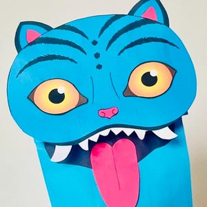 May include: A blue cat puppet with large yellow eyes, a pink nose, and a pink tongue sticking out. The puppet has white teeth and black stripes on its head. The ears are blue with pink inner details.