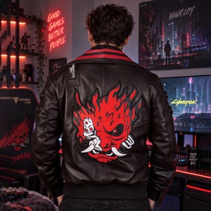 May include: A black leather jacket featuring a large, vibrant red and white graphic of a stylized oni mask on the back. The jacket includes a red collar trim, a small black tag with white text on the shoulder, and ribbed cuffs at the waist.
