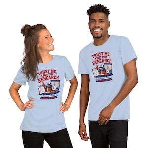 Könnte beinhalten: Ein hellblaues T-Shirt mit einer Grafik eines Footballs, der eine Sonnenbrille und einen Schaumstofffinger trägt, sowie dem in Rot und Blau gedruckten Text "TRUST ME, I DID THE RESEARCH" und "FANTASY FOOTBALL DRAFT DAY" auf der Vorderseite.
