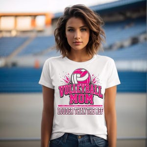 May include: White t-shirt with a pink volleyball graphic and the words "Volleyball Mom" above "Louder Than The Ref" in pink. The shirt is worn by a person in front of a stadium background.