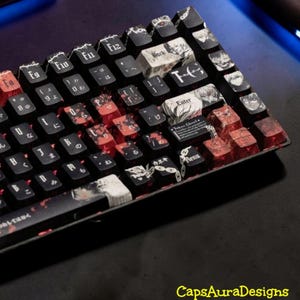 Könnte beinhalten: Eine mechanische Tastatur mit individuellen schwarzen Tastenkappen, die rote und weiße grafische Illustrationen wie Anime-Porträts, Ketten und japanische Schriftzeichen aufweisen. Die Tasten sind mit englischen Buchstaben und japanischen Zeichen beschriftet.
