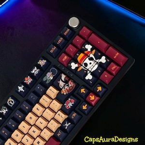 May include: A mechanical keyboard featuring a dark frame, a silver rotary knob, and custom keycaps. The keycaps display various pirate-themed skull graphics, orange lettering, and a large central skull icon. The text CapsAuraDesigns is visible.