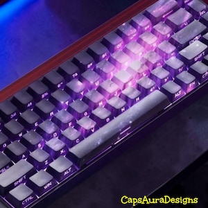 May include: A mechanical keyboard featuring black keycaps with a translucent, cloudy white marble-like pattern. The keys are illuminated by a vibrant purple backlight that shines through the letters and symbols. The text CapsAuraDesigns is visible.