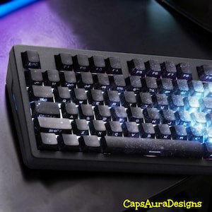 Könnte beinhalten: Eine schwarze mechanische Tastatur mit benutzerdefinierten Tastenkappen in einem dunklen Weltraum-Galaxie-Design, weißer Beschriftung und integrierter Hintergrundbeleuchtung. Der Text CapsAuraDesigns ist unten rechts gelb zu sehen.