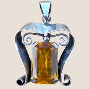 Puede incluir: Colgante de plata con una gran gema rectangular de color ámbar. El colgante tiene un diseño decorativo en espiral alrededor de la piedra y un lazo en la parte superior para una cadena.