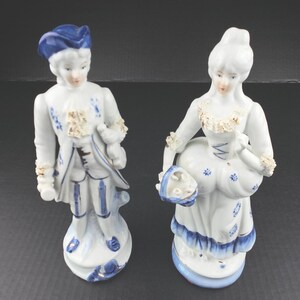 May include: Two porcelain figurines depicting a pair in historical attire. The male figure wears a blue hat and coat with white accents. The female figure wears a white dress with blue floral patterns, a ruffled collar, and carries a small basket.