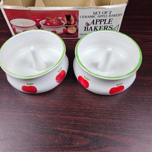 May include: A set of two white ceramic apple bakers featuring a central vertical spike and red apple illustrations on the exterior. The rim is accented with a thin green line. The original cardboard box is visible, labeled Set of 2 Ceramic Apple Bakers.