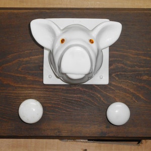 May include: A dark brown wooden wall mounted coat rack with a white ceramic pig head and four white ceramic knobs.