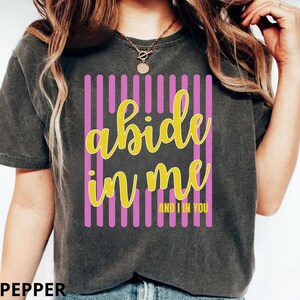 Puede incluir: Una camiseta de manga corta gris carbón con un diseño gráfico de rayas verticales rosas. El texto "abide in me" está impreso en letra cursiva amarilla grande, con el texto amarillo más pequeño "AND I IN YOU" debajo.