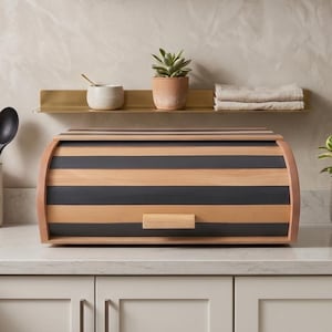 Puede incluir: Una panera rectangular con tapa enrollable que presenta rayas horizontales alternas de madera clara natural y gris carbón oscuro. El recipiente tiene un sencillo tirador de madera centrado en el panel frontal para abrir la tapa curva.