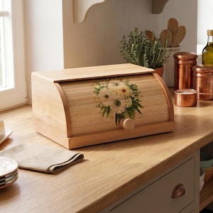 Peut inclure: Une boîte à pain en bois clair avec un couvercle coulissant orné d'un motif floral composé de fleurs blanches et de feuilles vertes. La boîte à pain possède un bouton rond en bois sur le devant et est posée sur un plan de travail en bois.