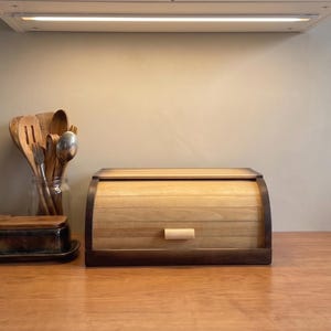May include: A rectangular wooden bread box with a roll-top lid featuring a light wood finish on the curved front and dark wood trim on the sides. The unit includes a small, light-colored wooden handle centered on the front panel for opening.