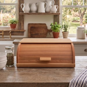 May include: A light brown wooden bread box with a roll-top lid and a small rectangular handle, positioned on a wooden kitchen counter. The bread box features horizontal slats on the top and a smooth, curved front panel made of natural wood.