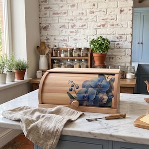 Puede incluir: Una panera de madera de color claro con tapa enrollable que presenta un estampado floral de orquídeas azules. La panera tiene un pequeño pomo redondo de madera para abrirla y está colocada sobre una encimera de mármol blanco.