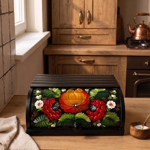 Puede incluir: Una panera de madera negra con puerta enrollable que presenta un diseño floral pintado a mano. La obra muestra una gran flor naranja central rodeada de racimos de bayas rojas, flores blancas y hojas verdes sobre un fondo oscuro.