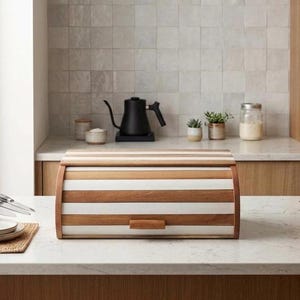 May include: A rectangular bread bin featuring alternating horizontal stripes of natural brown wood and white material, equipped with a roll-top lid and a small wooden handle, positioned on a white countertop next to a stack of plates.