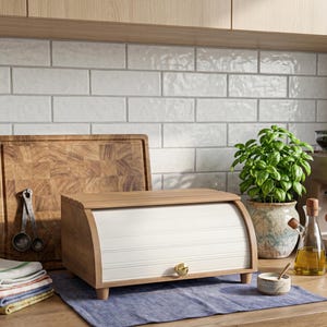 May include: A wooden bread bin with a roll-top door and a gold knob sits on a blue cloth. A wooden chopping board and measuring spoons are behind it. A small potted plant and a bottle of oil are also on the counter.