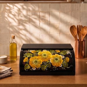 Peut inclure: Une boîte à pain en bois noir avec une porte coulissante ornée d'un motif floral peint à la main. L'illustration présente de grandes fleurs jaunes éclatantes avec des feuilles vertes et de petites fleurs blanches sur fond sombre.