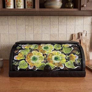 May include: A dark brown wooden bread box with a roll-top door featuring a hand-painted floral design. The artwork displays large yellow and green flowers with white petals and green leaves against a black background, centered with a round knob.