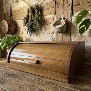 Puede incluir: Una panera rectangular de madera con una tapa enrollable de listones curvos y un pequeño tirador horizontal. El recipiente presenta un acabado de veta de madera de color marrón medio, una base plana y paneles laterales sólidos.