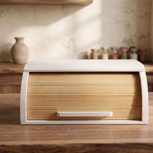 Peut inclure: Un coffre à pain en bois clair avec un dessus blanc et des côtés incurvés. Le devant présente des lattes horizontales en bois et une petite poignée rectangulaire. Le coffre à pain est posé sur une surface en bois.