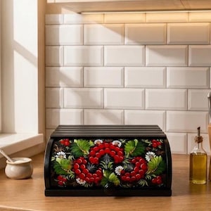 Puede incluir: Una panera de madera negra con tapa enrollable que presenta un diseño pintado a mano de bayas rojas, hojas verdes y pequeñas flores blancas. El panel frontal incluye un pequeño pomo negro para abrir la tapa con un motivo floral.