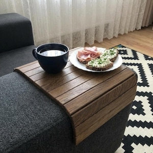 Könnte beinhalten: Ein hölzernes Sofatablett in warmbraunem Finish, das eine dunkelblaue Tasse und einen weißen Teller mit Essen hält. Das Tablett befindet sich auf einer grauen Sofalehne. Der Teller enthält zwei belegte Brote und Aufschnitt.