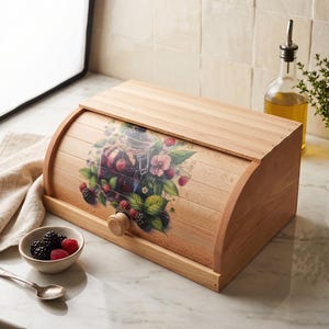 May include: A light-colored wooden bread box with a roll-top door featuring a printed graphic of a glass jar filled with blackberries, raspberries, and green leaves. The door has a small round wooden knob for opening and closing the container.