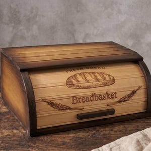 May include: A rectangular wooden bread box with a roll-top lid featuring a dark brown stained frame and a lighter natural wood front panel. The front is engraved with a loaf of bread, wheat stalks, and the text "FRESH BREAD" and "Breadbasket".