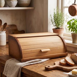 Op de afbeelding: Een houten broodtrommel met een roldeksel en een klein handvat. De trommel is gemaakt van lichtgekleurd hout en staat op een houten aanrecht. Gesneden brood en een snijplank zijn in de buurt.