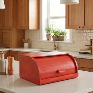 May include: A rectangular red wooden bread box with a roll-top lid and a small handle. The bread box features horizontal slats across the curved front and top surface, providing a functional storage solution for kitchen countertops.