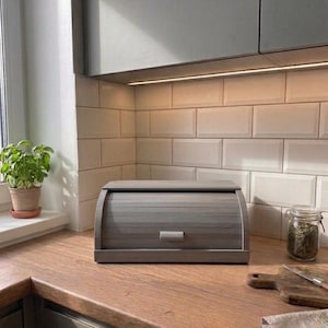 Könnte beinhalten: Ein rechteckiger Brotkasten aus Holz mit einem gewölbten Rolldeckel und einem kleinen horizontalen Griff. Der Brotkasten hat eine glatte, matte graue Oberfläche und ein Lamellendesign auf der Vorderseite für eine praktische Aufbewahrung.