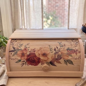 Può includere: Un portapane in legno chiaro con un design floreale con fiori rossi, arancioni e marroni. Il portapane ha una parte superiore curva e un pomello in legno. Lo sfondo comprende una finestra, utensili da cucina e una burriera.
