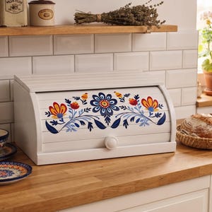 May include: A white wooden bread bin with a roll-top lid featuring a hand-painted floral design in blue, red, and yellow. The rectangular container has a small round knob on the front panel and a smooth finish, providing a storage solution.
