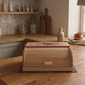 Puede incluir: Una panera rectangular de madera con tapa enrollable que incluye un pequeño tirador de madera y un diseño floral decorativo en el panel superior. La panera está fabricada en madera clara con listones horizontales en la puerta frontal.