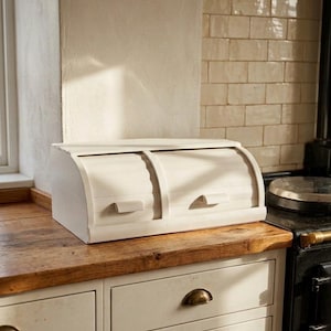 May include: A cream-coloured, roll-top bread bin with two pull-down handles sits on a wooden countertop. The kitchen cabinet below has cream drawers with brass handles. The background includes a cream wall and a black stove.