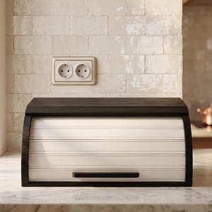 May include: A black wooden bread bin with a roll-top door and a white, slatted front. It is positioned on a marble countertop in a kitchen. An electrical outlet is visible on the wall behind the bread bin. The bread bin is approximately 40 cm wide.