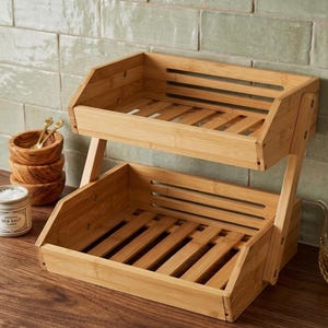 Op de afbeelding: Een bamboe plank met twee niveaus, met lattenbodems en schuine zijkanten. De plank in natuurlijke houttinten wordt ondersteund door een frame en is ontworpen voor opslag. Kleine houten kommen en een pot zeezout zijn zichtbaar op de achtergrond.