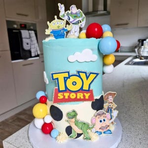 May include: A two-tiered cake decorated with Toy Story characters. The top tier is light blue with white cloud accents and the Toy Story logo. The bottom tier is white with black spots and character cutouts. Colourful ball decorations adorn the sides.