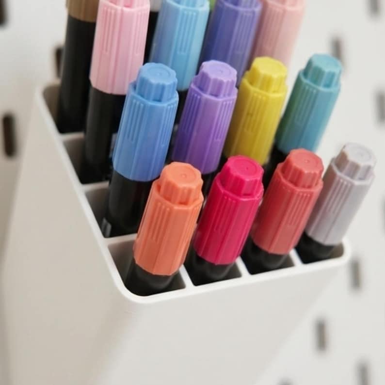 Peut inclure: Un organisateur en plastique blanc contenant une collection de marqueurs aux corps noirs et aux capuchons color&eacute;s dans des tons de rose, bleu, violet, jaune et gris. Les marqueurs sont dispos&eacute;s en grille dans le r&eacute;cipient rectangulaire.