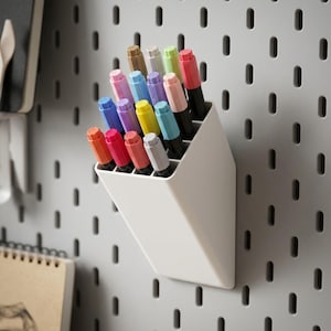Peut inclure: Un organisateur mural en plastique blanc avec un dessus inclin&eacute;, contenant une collection de quinze feutres de diff&eacute;rentes couleurs, dont rouge, rose, bleu, jaune, violet et vert, fix&eacute; sur un panneau perfor&eacute; gris &agrave; fentes verticales.