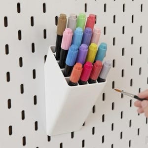 Peut inclure: Un r&eacute;cipient de rangement en plastique blanc dot&eacute; de plusieurs compartiments contenant une collection de marqueurs color&eacute;s dans des tons rose, bleu, jaune, vert et marron, fix&eacute; sur un panneau perfor&eacute; blanc avec des fentes verticales.