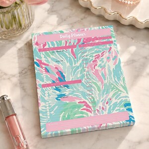 May include: A rectangular daily planner notepad featuring a colorful abstract pattern in shades of pink, teal, and blue. The page includes sections labeled Daily Planner, Work, Personal, Top 3 Priorities, To Do List, Daily Schedule, and Notes.