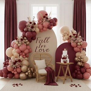 May include: A decorative event display featuring an arch backdrop with the text Fall In Love, surrounded by a balloon garland in shades of burgundy, dusty rose, and cream, accented with floral arrangements, a wicker chair, and a tiered cake.