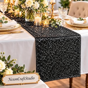 May include: A black sequin table runner draped over a white tablecloth. The runner features a dense, shimmering surface of small black sequins that reflect light. A gold-toned sign with the text NexonCraftStudio is in the foreground.