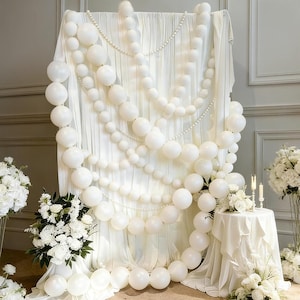 May include: A white fabric backdrop draped with multiple strands of varying sizes of white balloons and strings of white pearls. The display features a white-covered pedestal table with two lit candles and white floral arrangements on both sides.