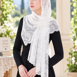 May include: A white lace head covering featuring a detailed floral pattern with scalloped edges. The fabric is draped over the head and shoulders, worn over a long-sleeved black dress. The lace has a delicate, semi-sheer texture throughout.
