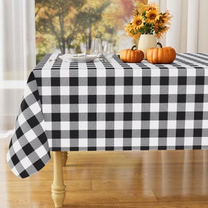 May include: A rectangular tablecloth featuring a black, white, and gray buffalo check pattern. The fabric drapes over a wooden table, creating a classic grid design. The surface is clean and smooth, showcasing the repeating square motif throughout.